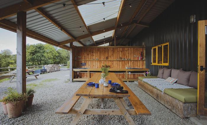 A large picnic table sits centre of an outdoor covered living space. There is a sofa to the right and washing up sinks in the background. Festoon ligh